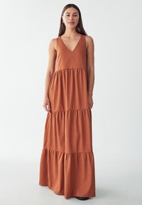Vestido largo en un cálido marrón, con escote en V, diseño sin mangas y dobladillo con volantes en capas. Hecho de una tela suave y fluida con una textura lisa.