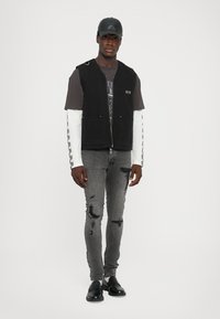 Vest negru fără mâneci, cu fermoar și buzunare frontale, purtat peste un tricou gri cu imprimeu, cu mâneci albe lungi și blugi gri uzati. Pantofi negri.