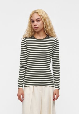 Femme aux longs cheveux blonds bouclés portant un haut à manches longues rayé horizontalement vert et blanc et un pantalon beige clair, debout devant un fond uni.