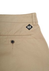 Beige bukser med en glat tekstur, udstyret med en baglomme med en mørk knap og et sort diamanformet logo med et "H" og "R."