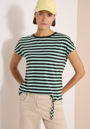 Femme portant une chemise à manches courtes rayée noir et bleu clair avec un lien sur le côté, pantalon beige et casquette jaune, debout les mains sur les hanches.