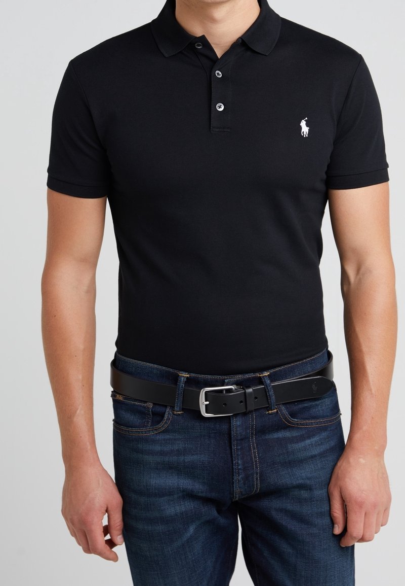 Polo noir en coton, avec une patte de boutonnage à trois boutons, des manches courtes et un petit logo blanc sur la poitrine gauche. Porté avec un jeans foncé.