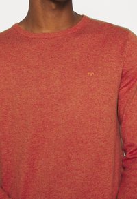 Pull rouge en maille avec un col rond, présentant un motif texturé subtil et un petit logo orange brodé sur la poitrine.