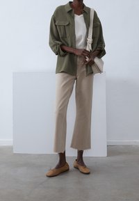 Olivově zelená košile na knoflíky se dvěma náprsními kapsami, bílý top s výstřihem do V, béžové zkrácené kalhoty s roztřepeným lemem a hnědé ploché boty.