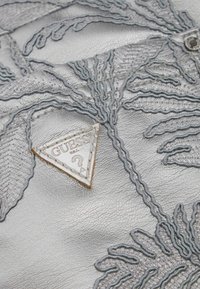 Pelle argentata con ricami grigi in rilievo a motivo foglia di palma. Presenta un'etichetta triangolare con logo Guess testurizzato.