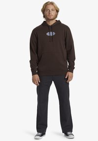 Sudadera marrón con bolsillo frontal y cordón, que presenta un logo azul. Combinada con pantalones grises a rayas y zapatillas en blanco y negro.