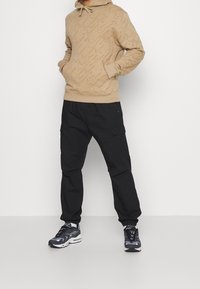 Tan huva sweatshirt med prägling av swoosh-mönster, ihopkopplad med svarta cargobyxor och grå-svarta sportskor.