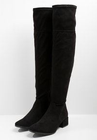 Bottes noires en daim hautes avec des talons blocs bas, des orteils ronds et une hauteur mi-mollet, se tenant debout sur une surface blanche.