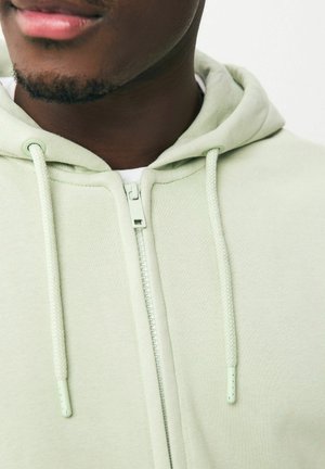 Felpa verde chiaro con zip, realizzata in tessuto testurizzato, dotata di cordini regolabili e di un dettaglio della cerniera liscia, che mette in risalto un design minimalista.
