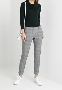 Pull à col cheminée vert foncé, pantalon taille haute à carreaux gris, et baskets blanches. La tenue comprend un petit sac à bandoulière blanc.