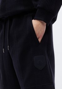 Μαύρο, ριμπ μπλουζάκι με κουκούλα και υφή υφάσματος· διαθέτει πλαϊνές τσέπες και ένα ανάγλυφο, μαύρο έμβλημα ραμμένο στο κάτω αριστερό μέρος.