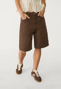 Mujer que lleva pantalones cortos marrones hasta la rodilla con bolsillos, combinados con zapatillas marrones y beige y una blusa de encaje crema.