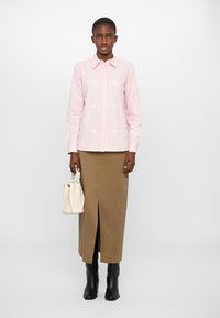 Camicia rosa a maniche lunghe con bottoni e ricami floreali bianchi, abbinata a una gonna beige fino al ginocchio con uno spacco frontale e stivali neri fino al ginocchio.