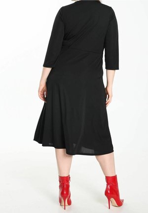 Kobieta ubrana w czarną sukienkę do kolan z rękawami do połowy ramienia oraz czerwone wysokie botki na obcasie, stojąca tyłem do widza.
