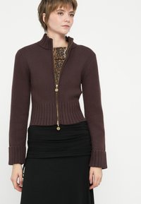 Žena s kratkom kosom nosi smeđu zip-up rebrastu džemper preko majice s leopard printom i naboranu crnu suknju, stojeći uz bijelu pozadinu.