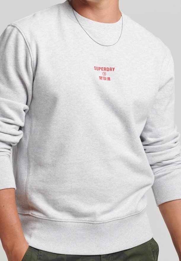 Sweat-shirt gris clair avec texte rouge "SUPERDRY" et petits caractères japonais sur la poitrine, porté avec les manches retroussées et un pantalon vert.