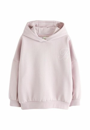 Jeune femme aux longs cheveux porte un sweat à capuche rose clair, un jean large bleu et des baskets blanches, debout devant un fond gris uni.