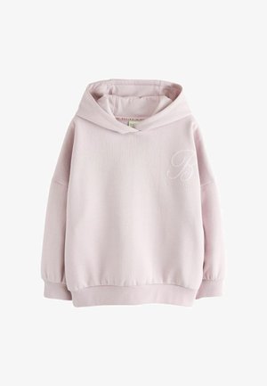 Młoda kobieta z długimi włosami nosi jasnoróżową bluzę z kapturem, niebieskie jeansy z szerokimi nogawkami oraz białe tenisówki, stoi na tle jednolitego szarego tła.