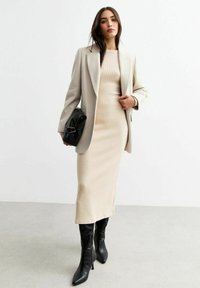 Abito in maglia a coste beige abbinato a un blazer grigio chiaro su misura e stivali neri con tacco a gamba alta, mentre tiene una borsa nera.