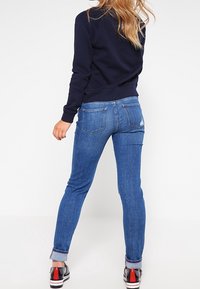 Woman wearing rolled-up blue jeans, black sneakers with red detail, and a dark navy sweatshirt, standing facing away on a plain background.