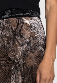 Lähikuva henkilön kädestä, joka lepää ruskeiden ja mustien käärmeennahkakuosisia samettihousuja sekä mustaa toppia vasten.