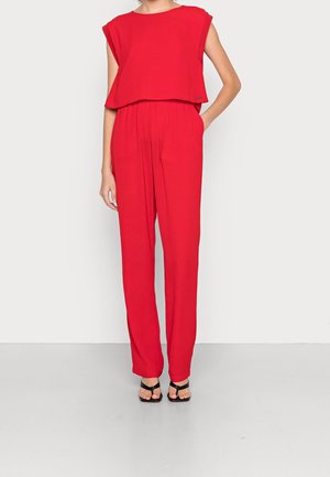 Czerwony, luźny kombinezon z krótkimi rękawami typu kaptur, elastycznym paskiem w talii i szerokimi nogawkami. Noszony z czarnymi sandałami na paski. Gładka faktura tkaniny.