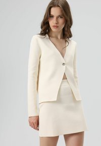 Blazer couleur crème avec un décolleté en V profond, un unique bouton argenté et des manches longues, associé à une mini-jupe taille haute assortie.