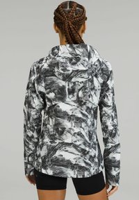 Sort og hvid mønstret jakke med hætte, struktureret stof, tætsiddende design og lange ærmer, med en buet kant bagpå.