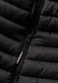 Chaqueta de abrigo negra con cierre frontal con cremallera y tirador de marca, textura acolchada y acabado de tela suave.