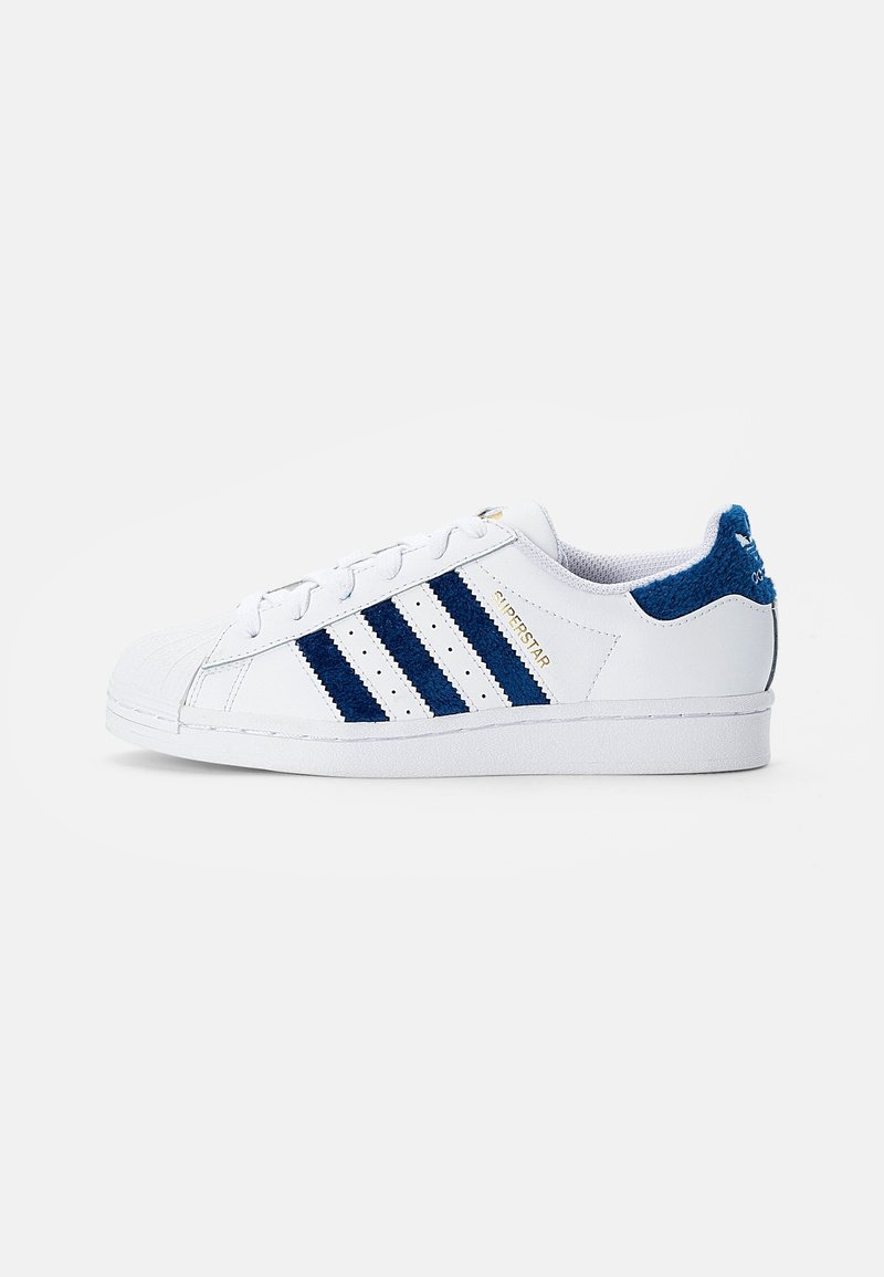 White sneaker featuring blue stripes and accents, textured back heel, rubber sole, and distinctive shell toe design.