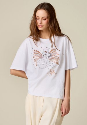 Jeune femme aux longs cheveux portant un t-shirt blanc oversize avec une illustration scintillante de fée avec des ailes et des cristaux décoratifs.