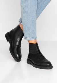 Černé kožené kotníkové boty s wingtip designem, šněrováním vpředu, prošívanými detaily a plochou gumovou podrážkou. Nosené s světle modrými zkrácenými džíny.