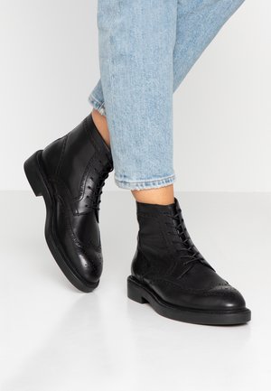 Mustat nahka nilkkurit, joissa on siipikuvio, nauhakiinnitys, tikkausyksityiskohtia ja litteä kumipohja. Yhdistetty kevyesti vaalean sinisiin farkkuihin, joissa on lyhennetty lahje.