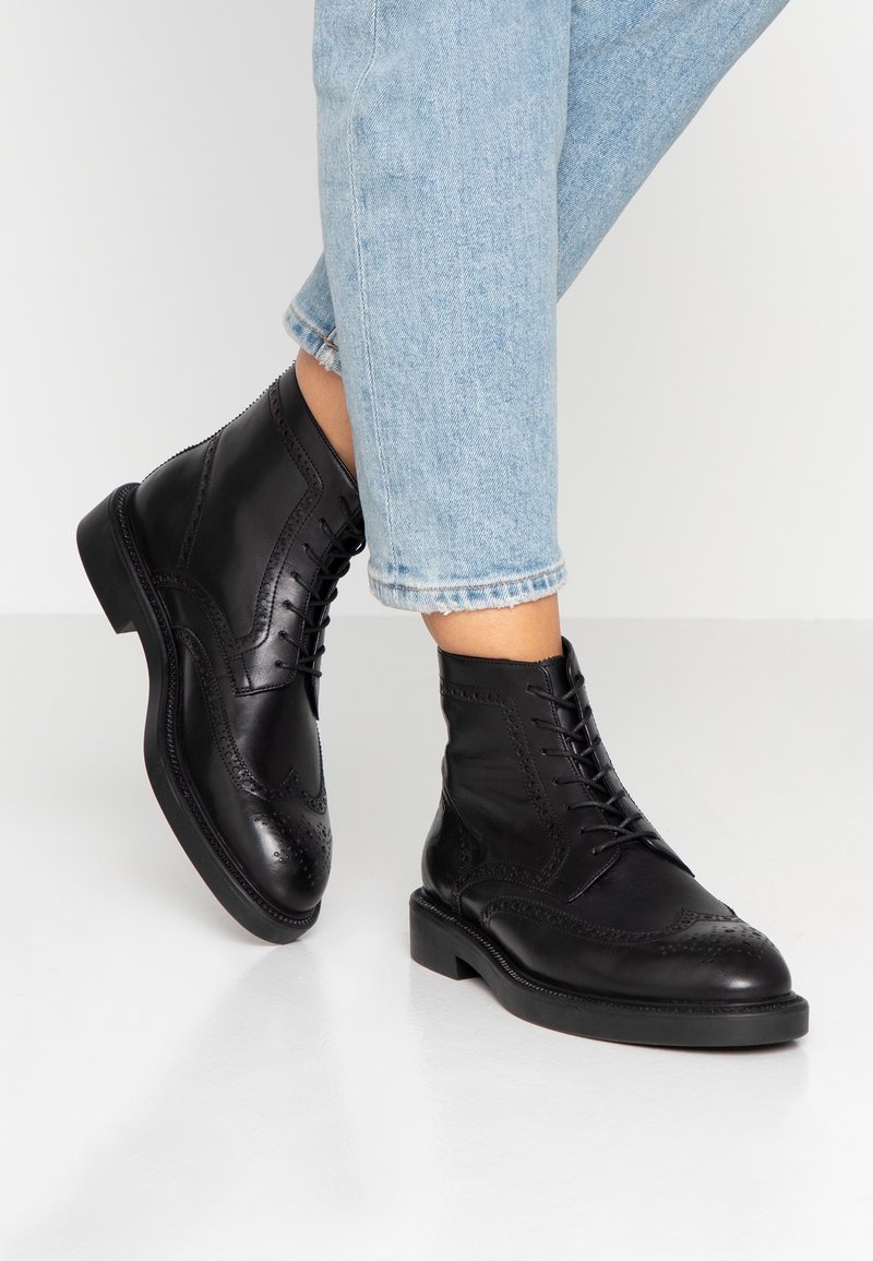 Černé kožené kotníkové boty s wingtip designem, šněrováním vpředu, prošívanými detaily a plochou gumovou podrážkou. Nosené s světle modrými zkrácenými džíny.