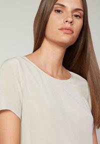 Kortärmad, rundhalsad topp i ljus beige tyg, med en slät textur och en ömtålig lyster, samt en subtil drapering vid axlarna.