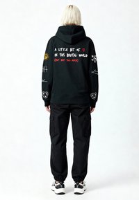 Felpa nera con cappuccio con testo bianco e rosso sulla schiena, con design grafici. Abbinata a pantaloni cargo neri e scarpe da ginnastica.