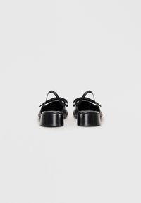 Črne slingback čevlje z gladkim usnjenim zaključkom, okroglo konico in nizko debelo peto. Detajl zankastega traku s srebrnimi okrasnimi elementi.