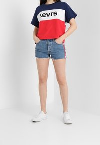 Červené, bílé a modré zkrácené tričko s logem "Levi's", v kombinaci se světle modrými džínovými šortkami s třásněmi a bílými teniskami.