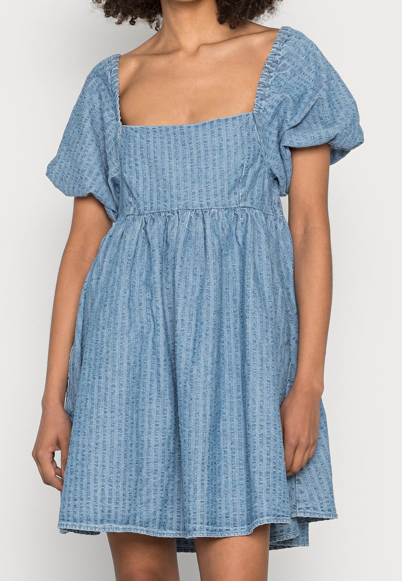 Robe en denim bleu avec un col carré, des manches bouffantes et une jupe plissée. Le tissu présente une texture subtile à rayures verticales.