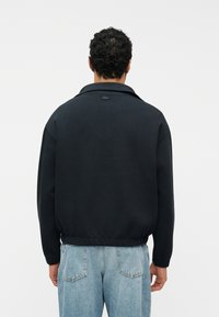 Άν δρας που φοράει μαύρο μακρυμάνικο μπουφάν και ανοιχτό μπλε τζιν, στέκεται γυρισμένος με την πλάτη σε λευκό απλό φόντο.