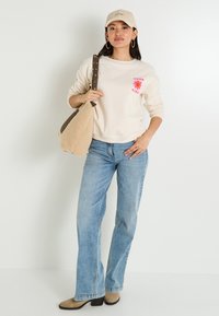 Jeune femme portant un sweat-shirt beige "Good Vibes", un jean bleu clair, une casquette beige, des boucles d'oreilles créoles, des bottes beiges et portant un sac à bandoulière texturé beige.