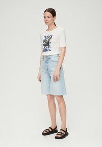 Witte korte mouwen crop top met een blauw en groen grafisch ontwerp, gecombineerd met lichtblauwe denim shorts met een rafelrand en zwarte sandalen.