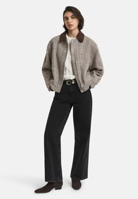 Bruine en grijze fleece bomberjack met een donkerbruine kraag, gedragen over een witte gebreide trui, gecombineerd met hoge zwarte jeans en laarzen.