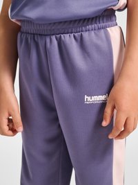 Child wearing purple and pink hummel Performance tracksuit pants with elastic waistband, hands relaxed at sides.