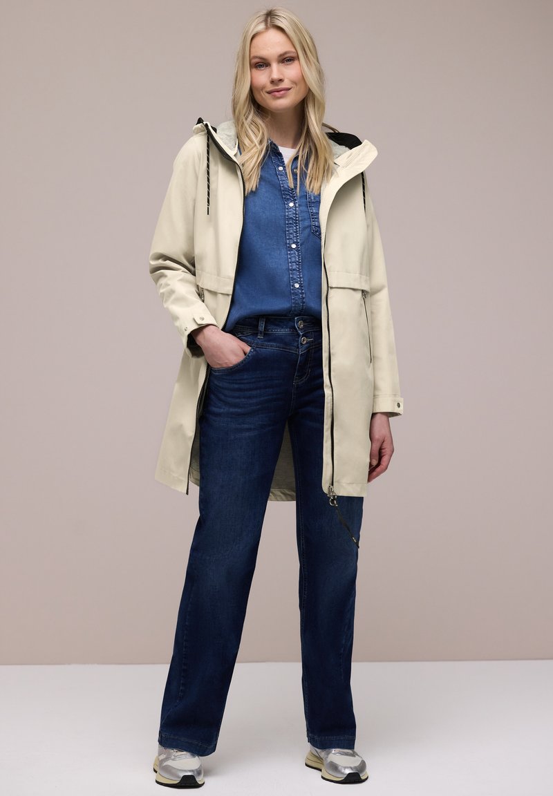 Femme blonde portant un imperméable à capuche beige sur une chemise en denim bleue et un jean, debout avec une main dans la poche devant un fond neutre.