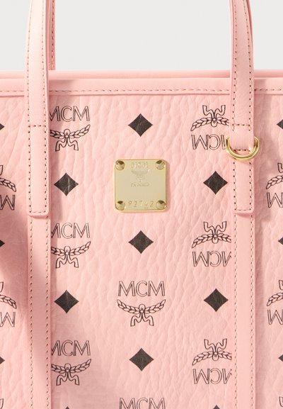 Różowa torebka skórzana o fakturowanej powierzchni z powtarzającym się czarnym wzorem logo MCM, złotymi akcentami i prostokątną tabliczką z nazwą.