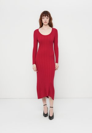 Red ribbed long-sleeve dress with a scoop neckline, fitted shape, knee-length hem, and side slit. Worn with black ankle strap heels.