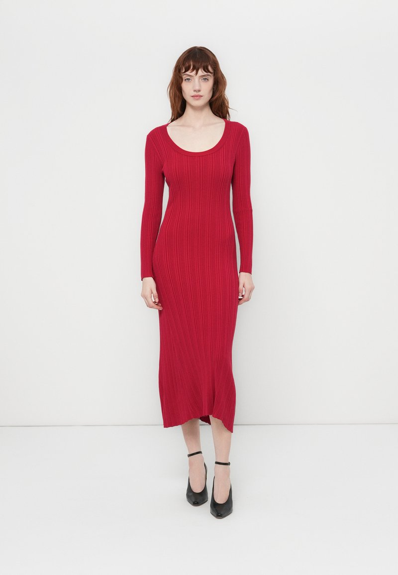 Robe longue à manches longues rouge côtelée avec un décolleté dégagé, silhouette ajustée, ourlet à mi-genoux et fente latérale. Portée avec des talons à lanières noirs.