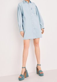 Vestito camicia in denim blu chiaro con maniche lunghe, chiusura con bottoni, tasca sul petto; abbinato a sandali con piattaforma color turchese e cinturini alla caviglia.