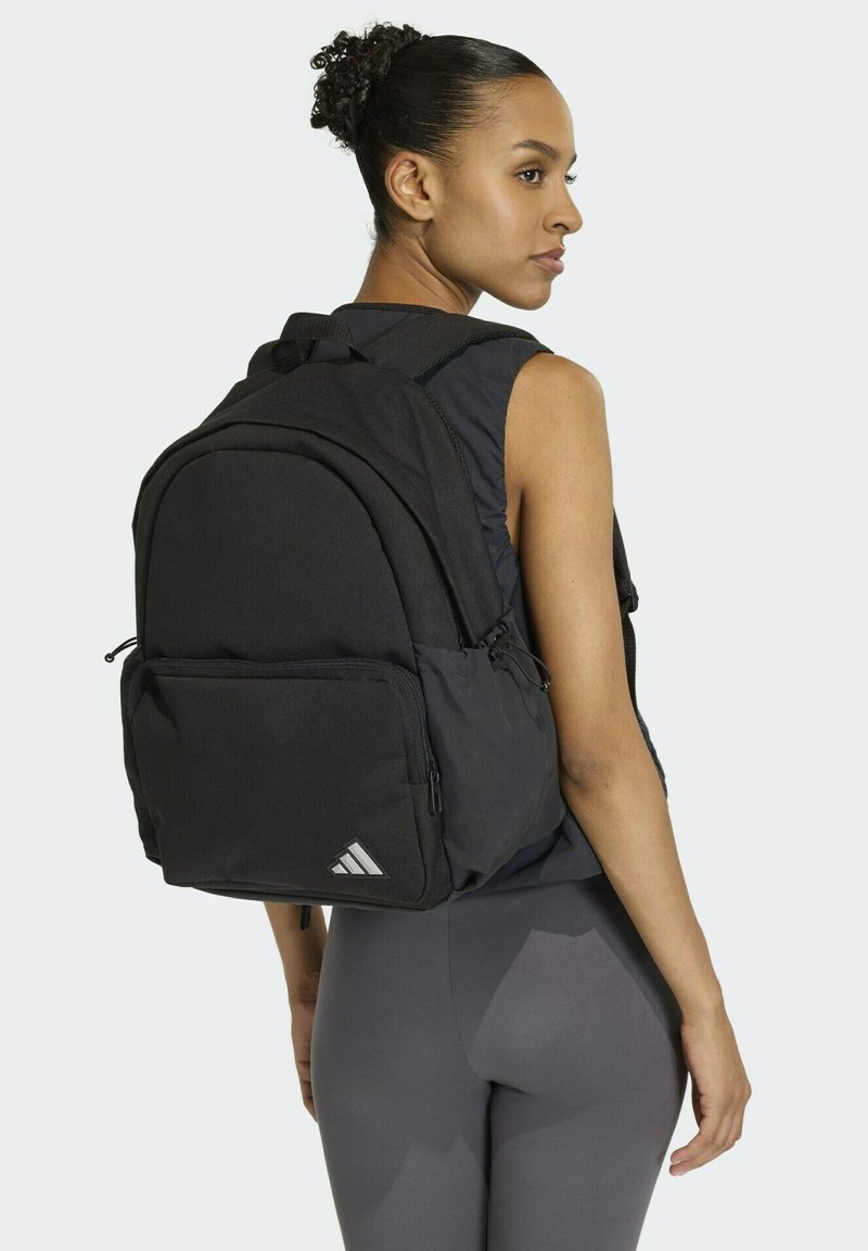 Mochila negra con correas acolchadas, un compartimento principal grande, bolsillo frontal con cremallera y detalles reflectantes en el logo, con un tejido texturizado.